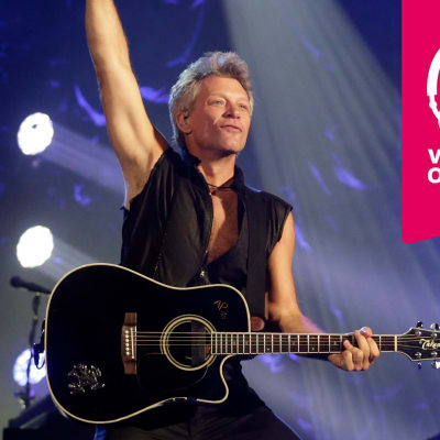 Jon Bon Jovi har en akustisk gitarr kring halsen och han sträcker höger arm rakt upp i luften.