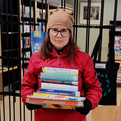 En kvinna står med en hög böcker i famnen inne i ett bibliotek.