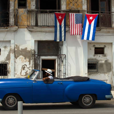 Vanha auto ohittaa taloa jossa roikkuu Yhdysvaltain ja Kuuban lippuja.