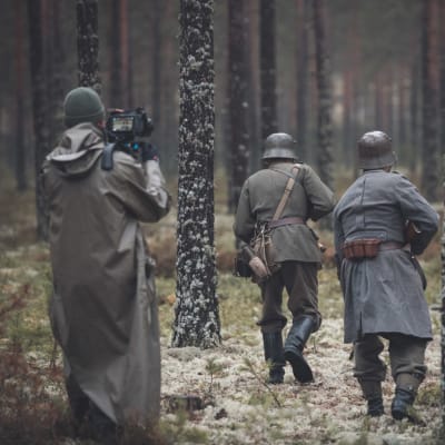Inspelning i skog, soldater (skådespelare) springer med ryggen vänd mot kameran mellan tallar. De har hjälmar och gevär.