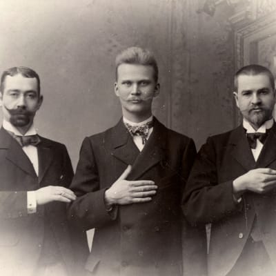 Kolme nuorta miestä seisovat vanhassa valokuvassa, esittäen käsillään viittomakielen Maamme Laulu viittomia.