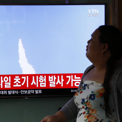 Televisioruutu, jossa näkyy ilmassa lentävän ohjuksen vana. Kuvan alareunassa on koreankielistä tekstiä. Televisioruudun oikealla puolella istuu nainen, joka katsoo kohti televisiota.