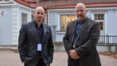 Två medelålders män i kavaj står ute på en gårdsplan framför ett ljusblått stenhus med rött tak.