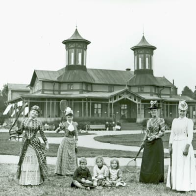 Fyra kvinnor i långa klänningar och fina hattar framför en utsmyckad träbyggnad. Mellan dem sitter tre barn i gräset. Kvinnorna håller alla i tennisracketar. Bilden är svartvit och från 1910.