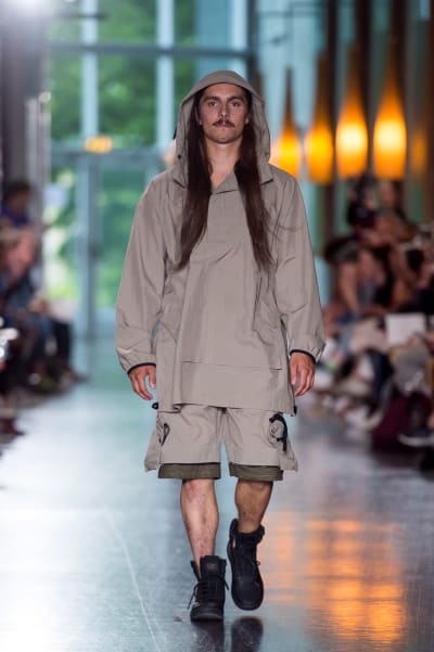 En manlig modell som går på catwalken. Han är iklädd beige shorts och en beige parkas.