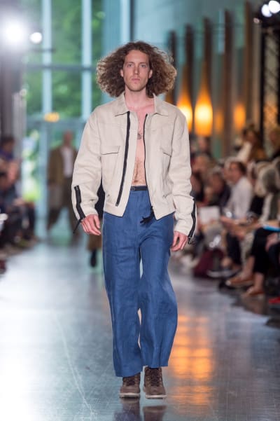En manlig modell som går på en catwalk. Han är klädd i blåa byxor och en beige jacka. 