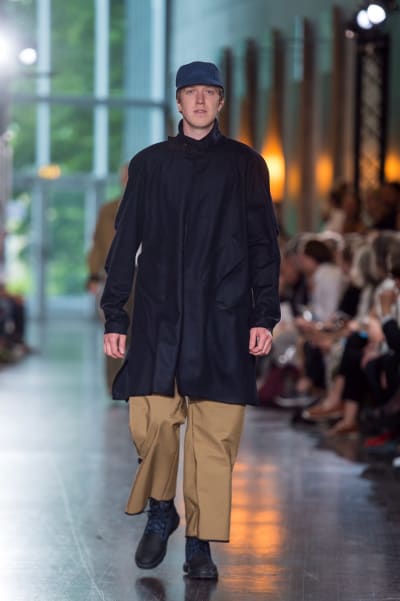 En manlig modell som går på en catwalk. Han är klädd i beige byxor, en mörkblå jacka och en mörkblå keps. 