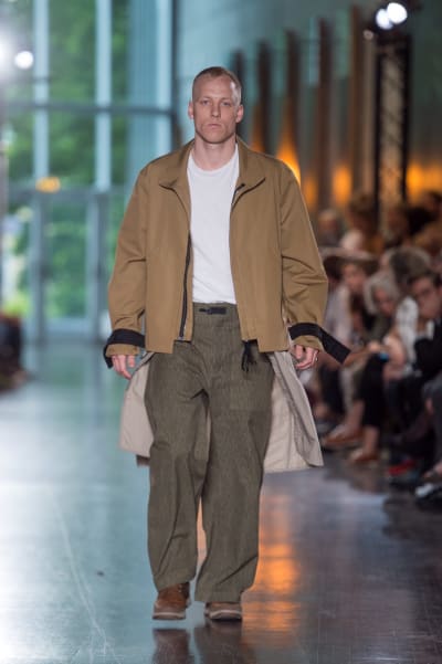 En manlig modell som går på en catwalk. Han är klädd i gråbruna byxor, en vit t-skjorta och en beige jacka. 