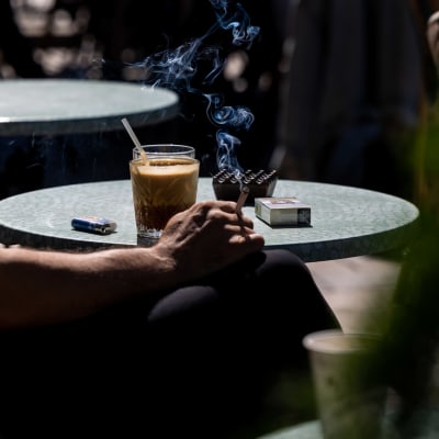 Anonyymi ihminen juo kahvia ja polttaa tupakkaa aurinkoisella terassilla. 