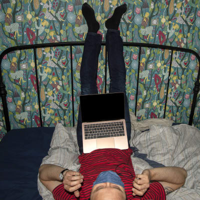 Kasvomaskiin pukeutunut henkilö makaa sängyllä kannettava tietokone sylissään.