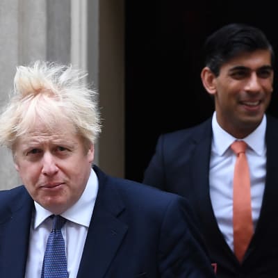 Boris Johnson tukka pystyssä. Taustalla Rishi Sunak.