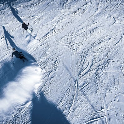 Aurinko paistaa, kun laskettelijat kiitävät alas lumen peittemää rinnettä.