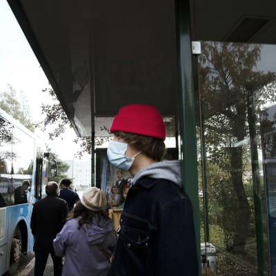 Kasvomaskiin pukeutunut nuori bussipysäkillä.