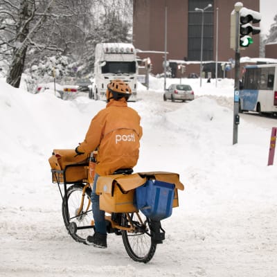 Postinjakaja ajaa polkupyörällä lumisessa katumaisemassa.