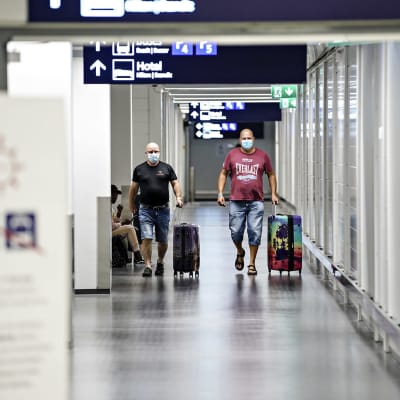 Två resenärer går med varsin kappsäck på Helsingfors-Vanda flygfält. Båda bär ansiktsskydd. 