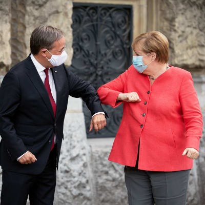 KAsvomaskeihin pukeutuneet Armin Laschet ja Angela Merkel kyynärtervehtivät toisiaan.