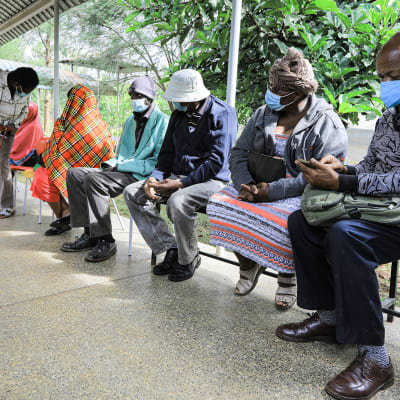 Ryhmä kenialaisia istuu ulkona odottamassa koronarokotetta.
