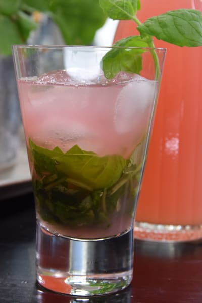 Ett glas med en rosa dryck, gröna örtblad och isbitar. I bakgrunden en flaska med mera rosa dryck.