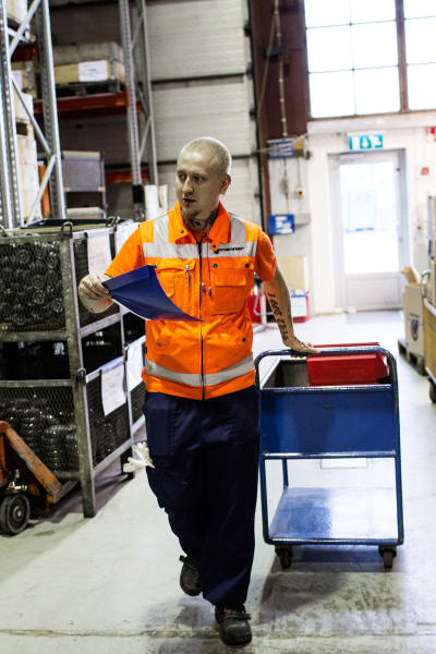 Sami Heinonen iklädd orange väst drar en kärra i fabrikshall.