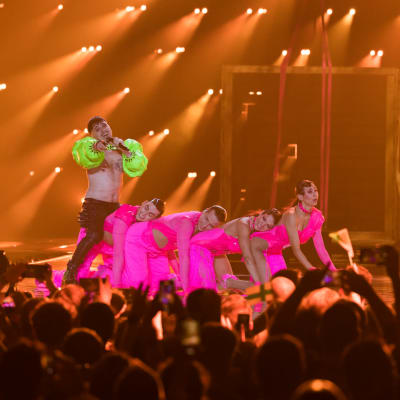 En grönklädd person och fyra personer klädda i ljusrött uppträder på en scen.