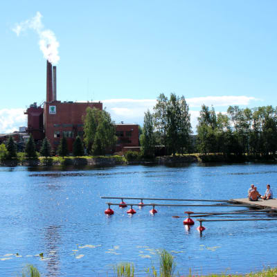 Kolme miestä istuu laiturilla kauniina kesäpäivänä ja Piesjoen toisella puolella näkyy vaneritehdas.