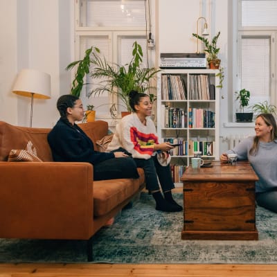 Kolme nuorta naista istumassa olohuoneessa, kaksi istuu sohvalla, yksi lattialla. Kuvan oikeassa laidassa on televisio, jonka ruudulla näkyy Yle Areena.
