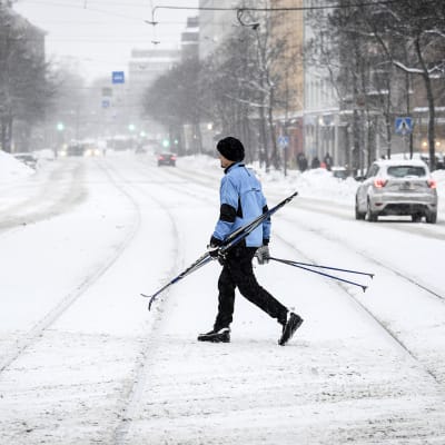 Suksia kantava mies ylittää lumipyryssä katua Helsingissä.