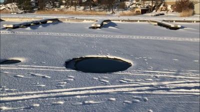 En stor rund öppen vak har bildats i isen mitt i vintern. I snön syns spår av snöskotrar och skidor, i bakgrunden bryggor vid stranden.