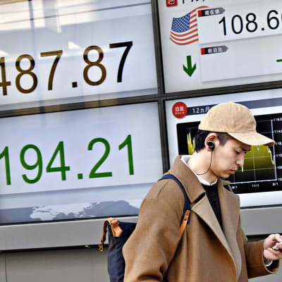 Nuori mies katselee älypuhelintaan Tokion kadulla 6. helmikuuta. Hänen takanaan näkyy laskevia pörssilukemia.