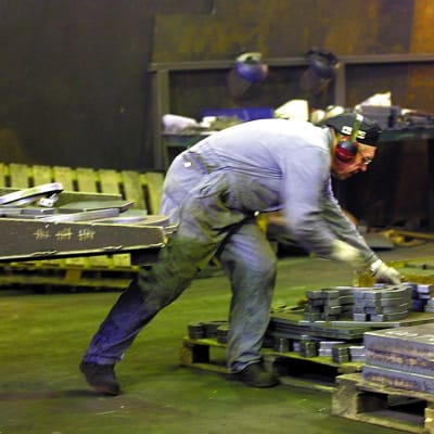 Nedre kroppen av person i overall lyfter metallföremål på fabriksgolv.