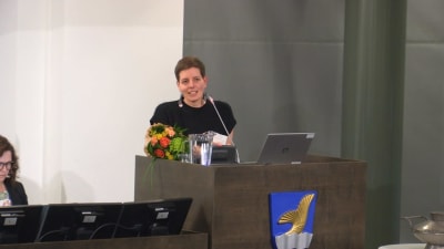 Åstrand håller tacktal i stadshuset podium, hon har en brombukett i handen. Vanda stads vapen pryder podiet.