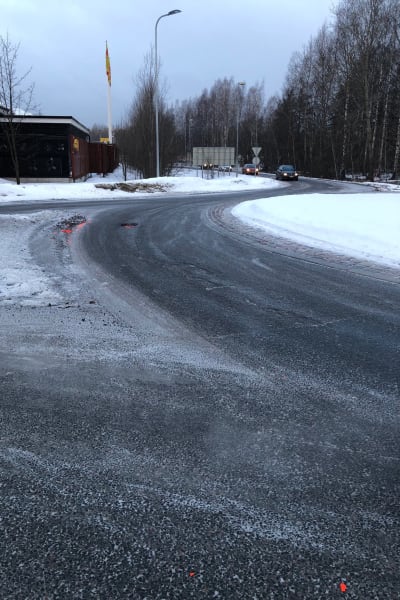 En rondell. Det är dag, det ligger snö på marken. I mitten av bilden finns en grop i asfalten.Det ligger också orangea plastbitar på asfalten.