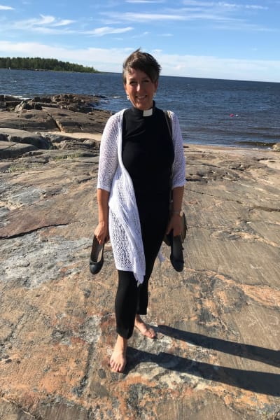 Prästen Patrica Strömbäck med skorna i handen på en klippa vid havet i Pörkenäs.