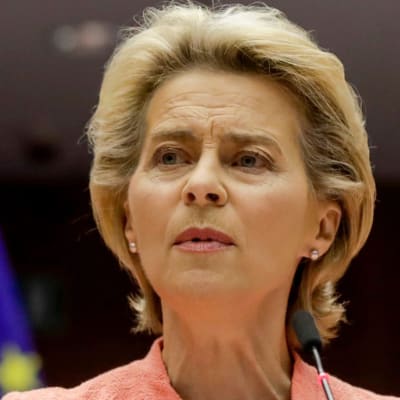 Ursula von der Leyen i närbild. Bakom henne syns en EU-flagga och en svart bakgrund. Von der Leyen talar i mikrofon och har händerna uppe, hon har en laxröd kavaj på sig.
