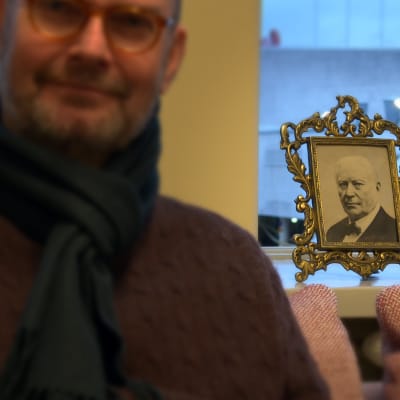 Porträtt på museichef Kai Kartio (suddig) med foto på Amos Andersson i bakgrunden (skarp).