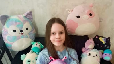 11-vuotias tyttö katsoo vienosto hymyillen kameraan. Hän istuu sohvalla ja pitää sylissään Squishmallows-pehmohahmoja. Myös hänen ympärillään on Squishmallows-hahmoja.