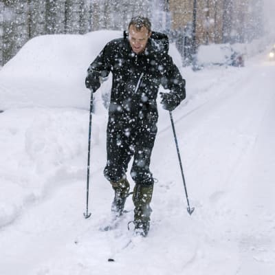 Lunta tuiskuttaa, mies kävelee lumisessa kaupungissa sauvojen kanssa.