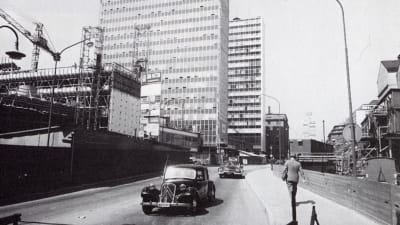 Ett svartvitt fotografi, bilar kör förbi platsen där Hötorgsskraporna i Stockholm håller på att byggas år 1959. 