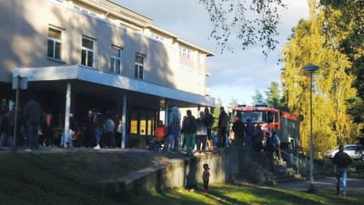 Människor står utanför en byggnad. En brandbil är parkerad bakom dem.