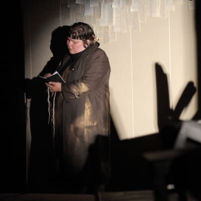 Kvinna klädd i svart läser en bok i en mörk och dramatisk miljö. Stillbild från en pjäs.