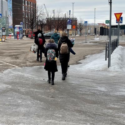 En grupp med personer, en familj, med ryggarna mot kameran, går bort. Det är i Helsingfors. Alla bär en ryggsäck eller väska..