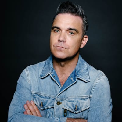 Robbie Williams seisoo tummaa taustaa vasten kädet puuskassa ja katsoo vakavana kameraan. Williamsilla on yllään farkkutakki.