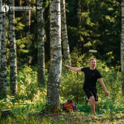En svartklädd man springer i skogen och ler. En frisbee flyger ovanför hans huvud. 