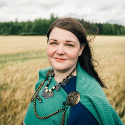 Personporträtt på kvinna med långt mörkt hår som står ute på ett sädesfält. Hon bär en grön sjal och smycken i brons.