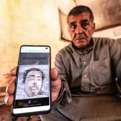 En sorgsen äldre man i gråbrun skjorta håller fram en mobil med en bild på en yngre man.