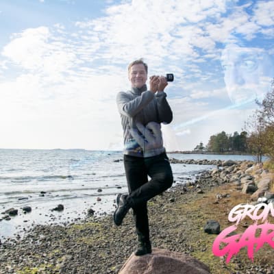 Carl-Kristian Rundman poserar på ett ben på strandsten och låtsas spela fläöjt med Yle-mikrofon. Ian Andersons ansikte bland molnen.