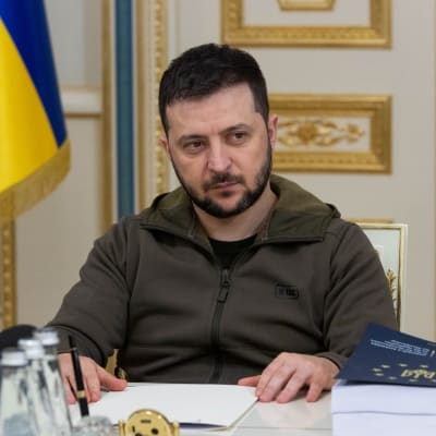 Volodymyr Zelenskyj sitter vid ett bord. Han tittar åt sidan. I bakgrunden Ukrainas flagga.