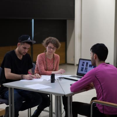 Tre personer vid ett bord, två unga män och en kvinna, sitter och jobbar med något. Ena mannen antecknar på ett papper, de två andra ser på. På borde syns en dator och på skärmen foton.
