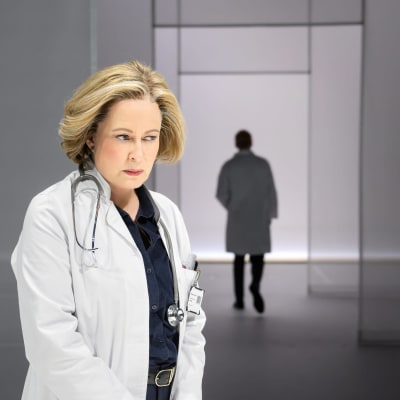 En kvinna i vit läkarrock står och sneglar efter siluetten av en person som avlägsnar sig. 