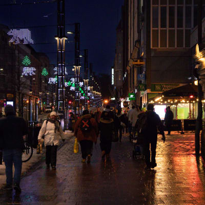Ihmisiä kävelemässä Hämeenkadulla. Tien yläpuolella on jouluvaloja, ja ympäristö on pimeä ja märkä.